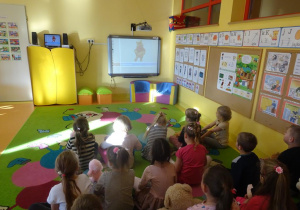 Grupa dzieci siedzi przed tablicą interaktywną, ogląda prezentację multimedialną na temat Misia pluszowego.